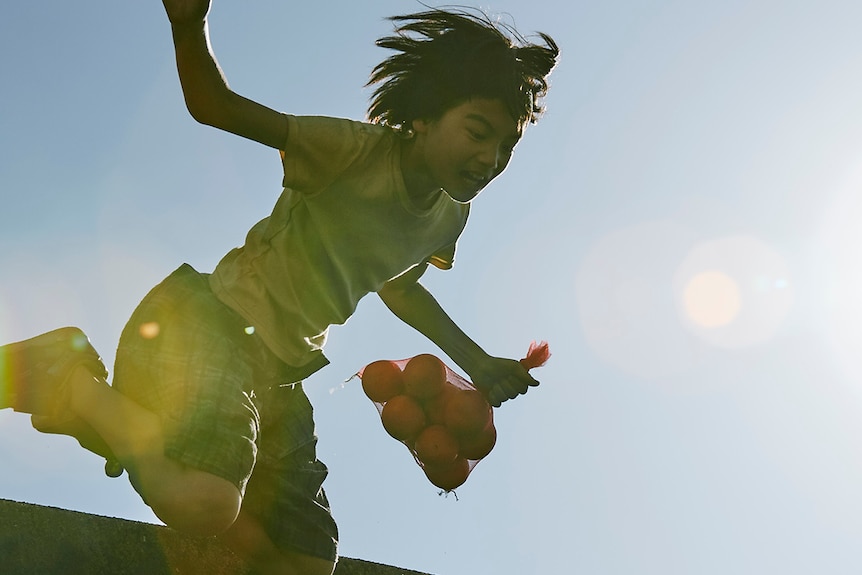 Colour still of Jyo Kairi in mid-air whilst jumping and holding bundle of oranges in 2018 film Shoplifters.