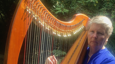 A woman stands holding her harp in front of some green trees.