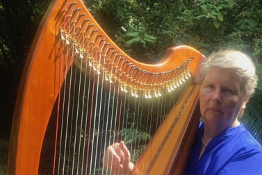 A woman stands holding her harp in front of some green trees.