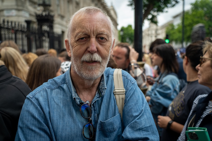 An older man with a grey beard and in a denim shirt stands in a group of people.
