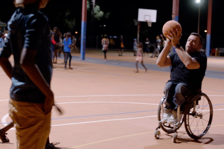 Mannin wheelchair on basketball court about to shoot the ball at the hoop. 