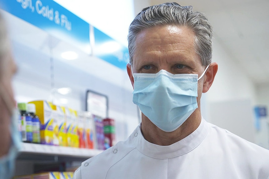 A man wearing a white chemist's uniform and a surgical mask.