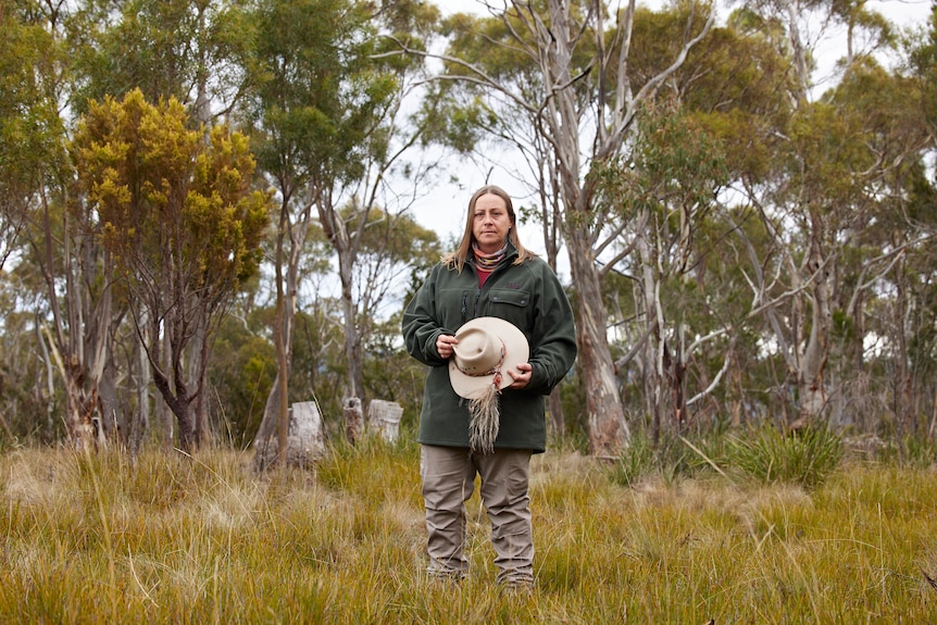 A woman in her early 40s in a big green coat and holding a white akubra hat standing in the bush