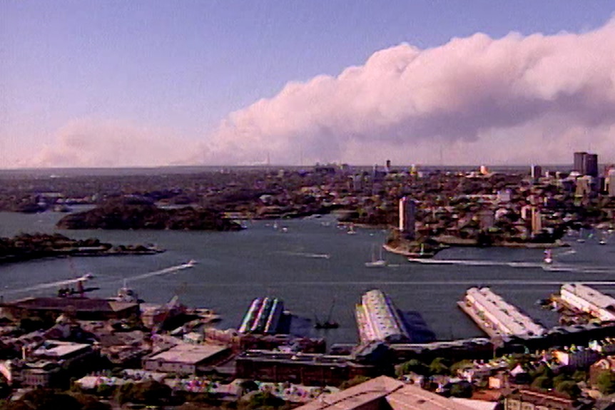 A large smoke plume rises above the sky above a city