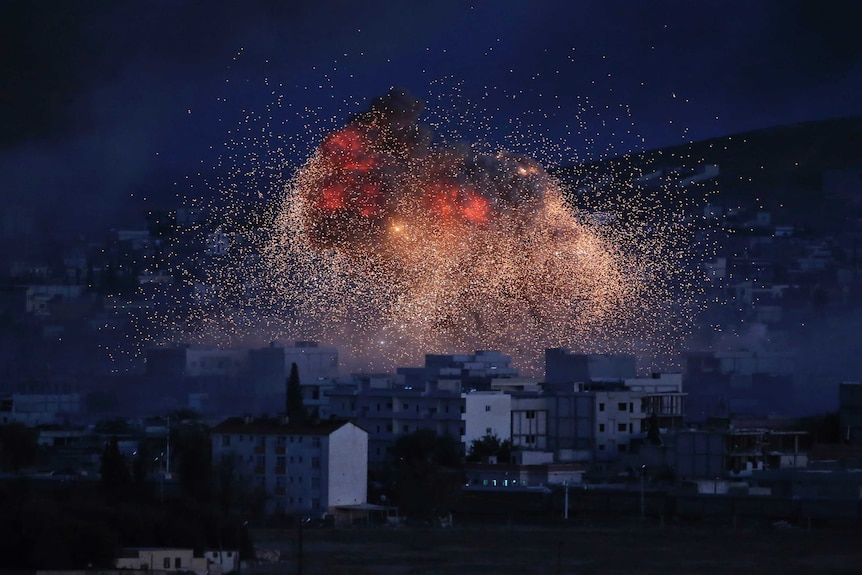 At dusk, a town is cast in blue light as an exploding bomb sprays bright red embers across it.