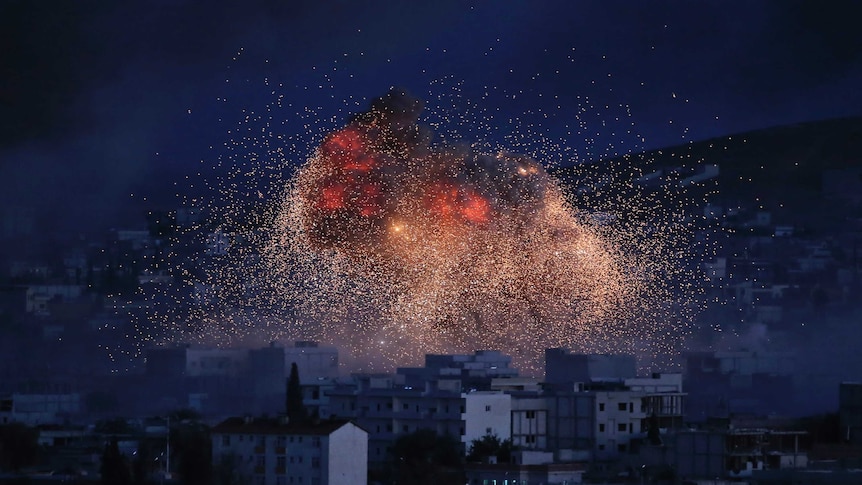 At dusk, a town is cast in blue light as an exploding bomb sprays bright red embers across it.
