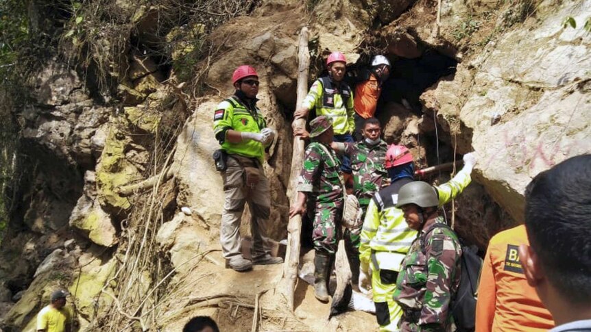 A group of Indonesian rescuers in high-vis are in a line up a hill at the entrance to a gold mine  carved into rock.