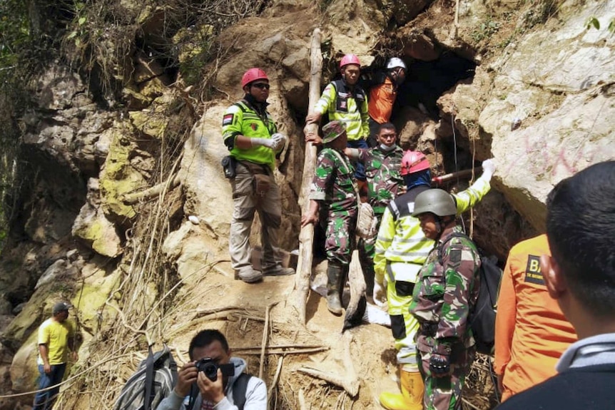 A group of Indonesian rescuers in high-vis are in a line up a hill at the entrance to a gold mine  carved into rock.