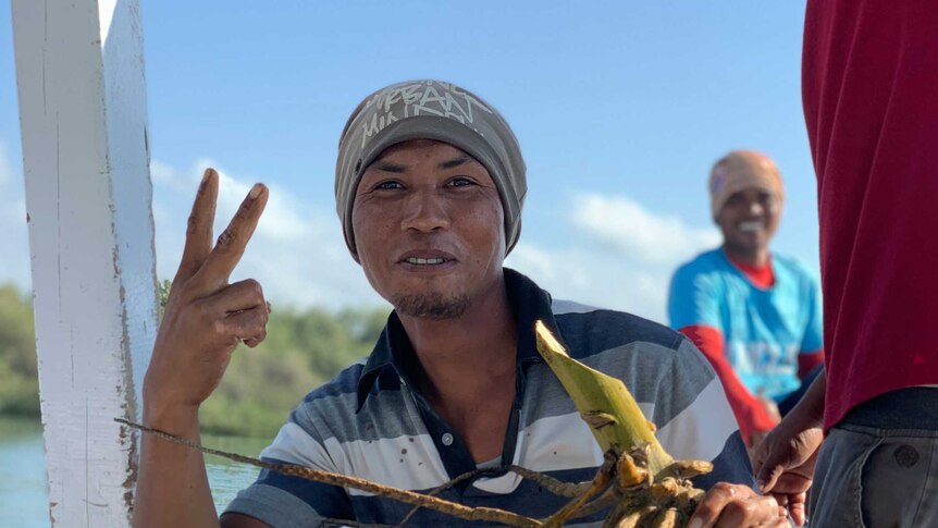 A man on a boat holds coconuts and gives the peace sign