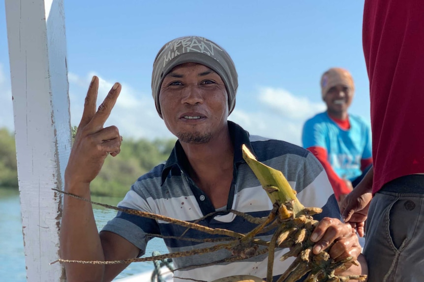 A man on a boat holds coconuts and gives the peace sign