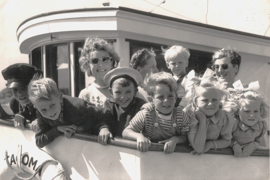 Black and white 1950s photo, seven smiley young children, three women looking out from top deck of white boat 