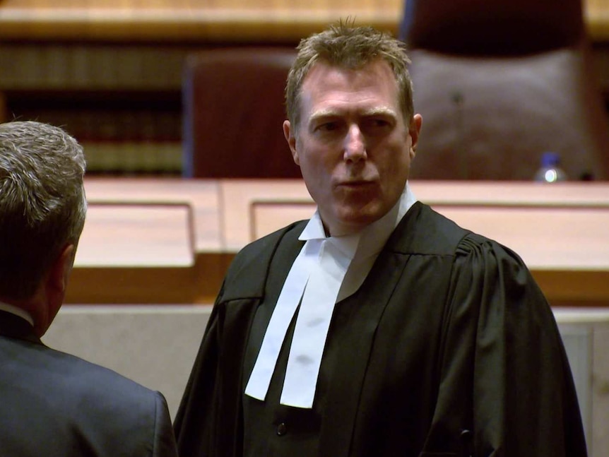 A man with short brown hair wearing a barristers robe with white collar looking to his left in front of a court bench