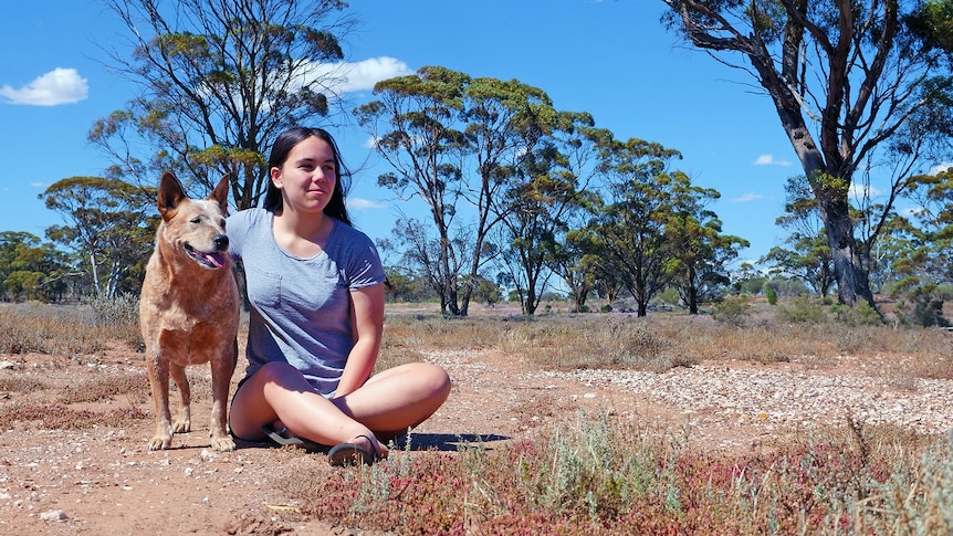 A young woman is sitting on the ground with her arm over a cattle dog. They're in grassy outback with a few gumtrees.