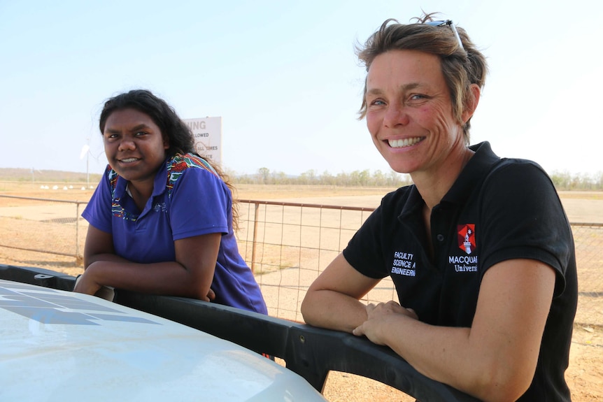 A student and Emilie Ens smile at the camera as they lean on a car bonnet