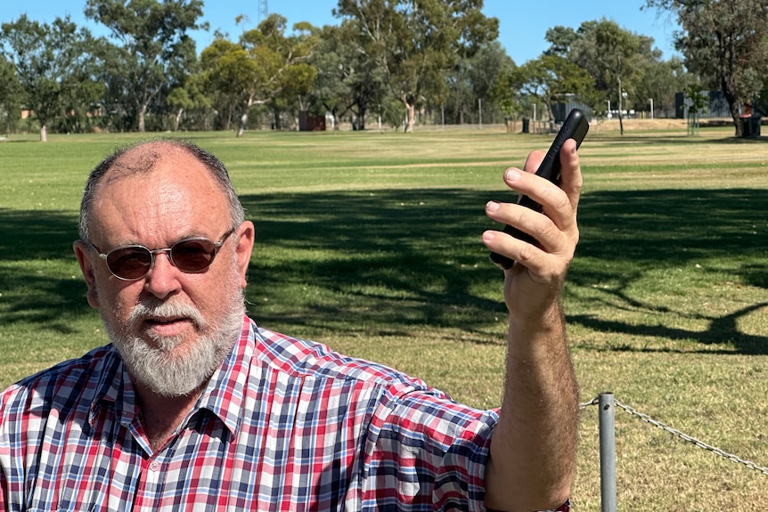 A close up of a man in his 60s with a beard and glasses, holding up his mobile phone trying to get reception in a grassy field