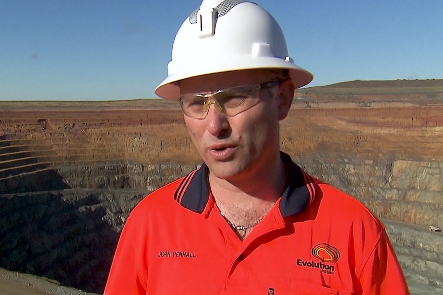 A man in a hard hat, safety glasses and high-vis stands talking in front of a mine pit.