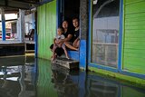 A young family - man, woman and child - sit on the step to their home, with floodwaters at their feet
