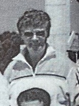 An old, black & white image of a man with dark, curly hair and wearing sunglasses standing behind and above another person