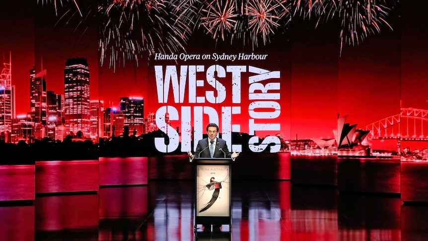 Lyndon Terracini stands at a podium in front of a projection of the Sydney skyline and the logo for West Side Story.