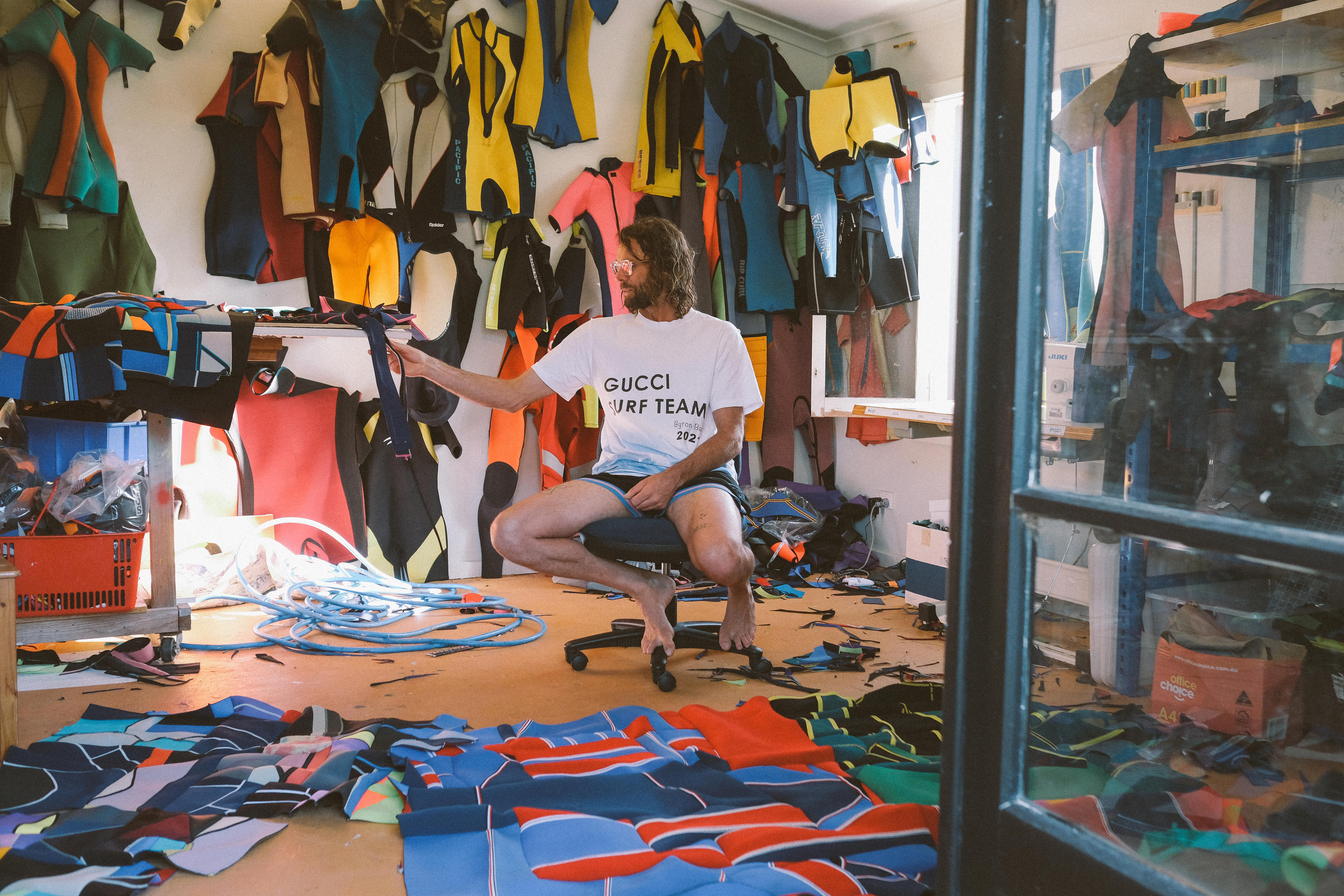 Artists sits in his studio surrounded by wetsuits and artworks in progress