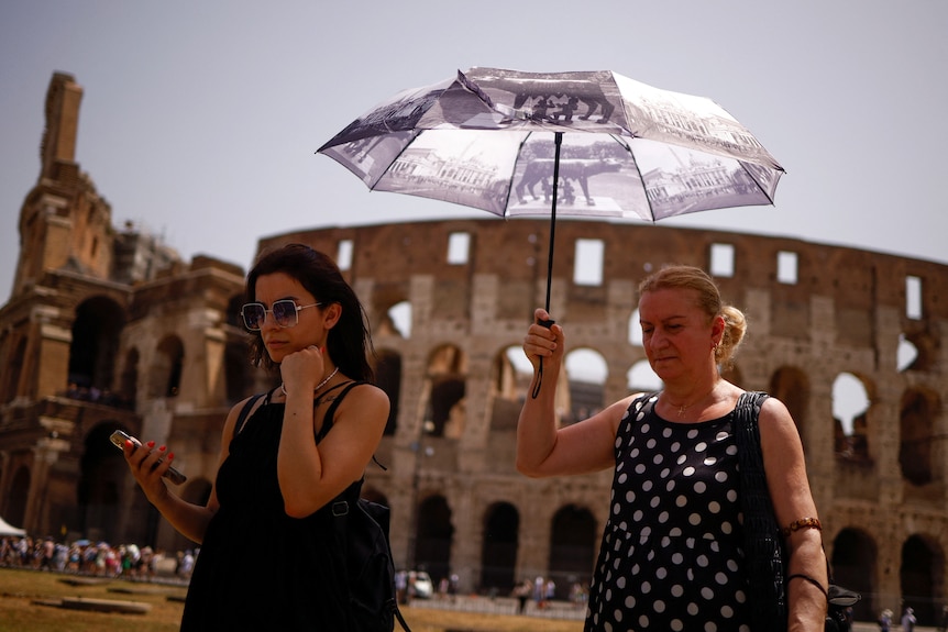 Two women carry umbrellas while walking past the Colosseum on a hot day