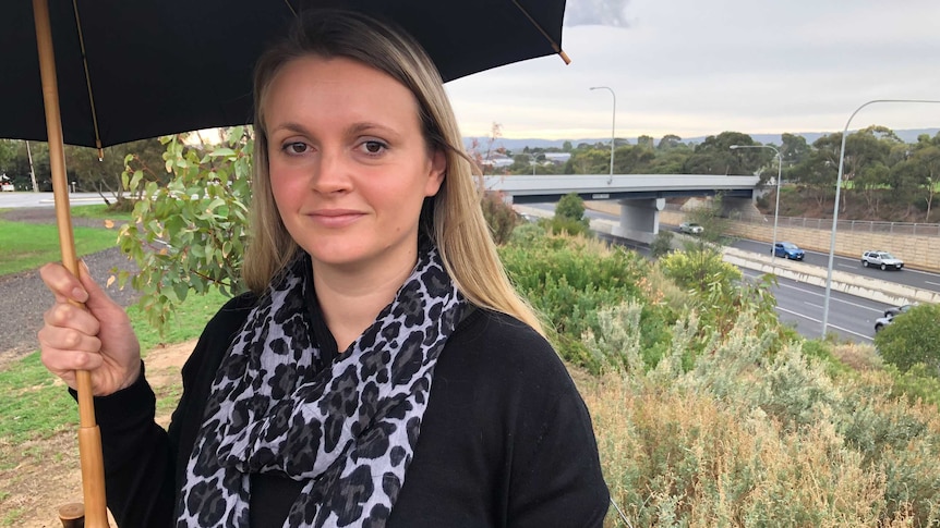 A woman holding an umbrella looks at the camera with a bridge in the background