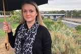 A woman holding an umbrella looks at the camera with a bridge in the background