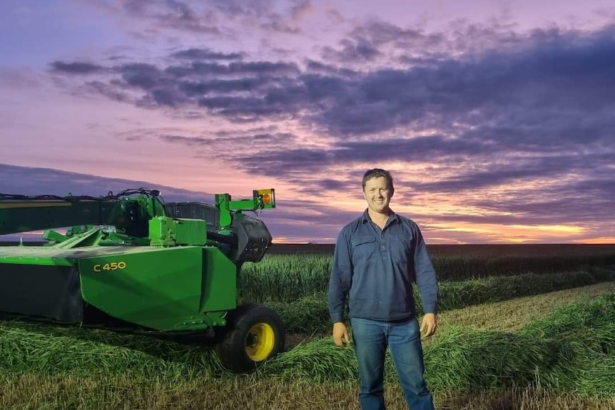 A smiling man in a shirt and blue jeans standing in a green paddock with machinery in background, sky almost purple at sunset.