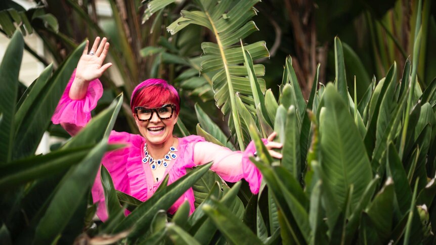 The comedian Cal Wilson in pink clothes and red hair and jewels amongst big green fronds of palms