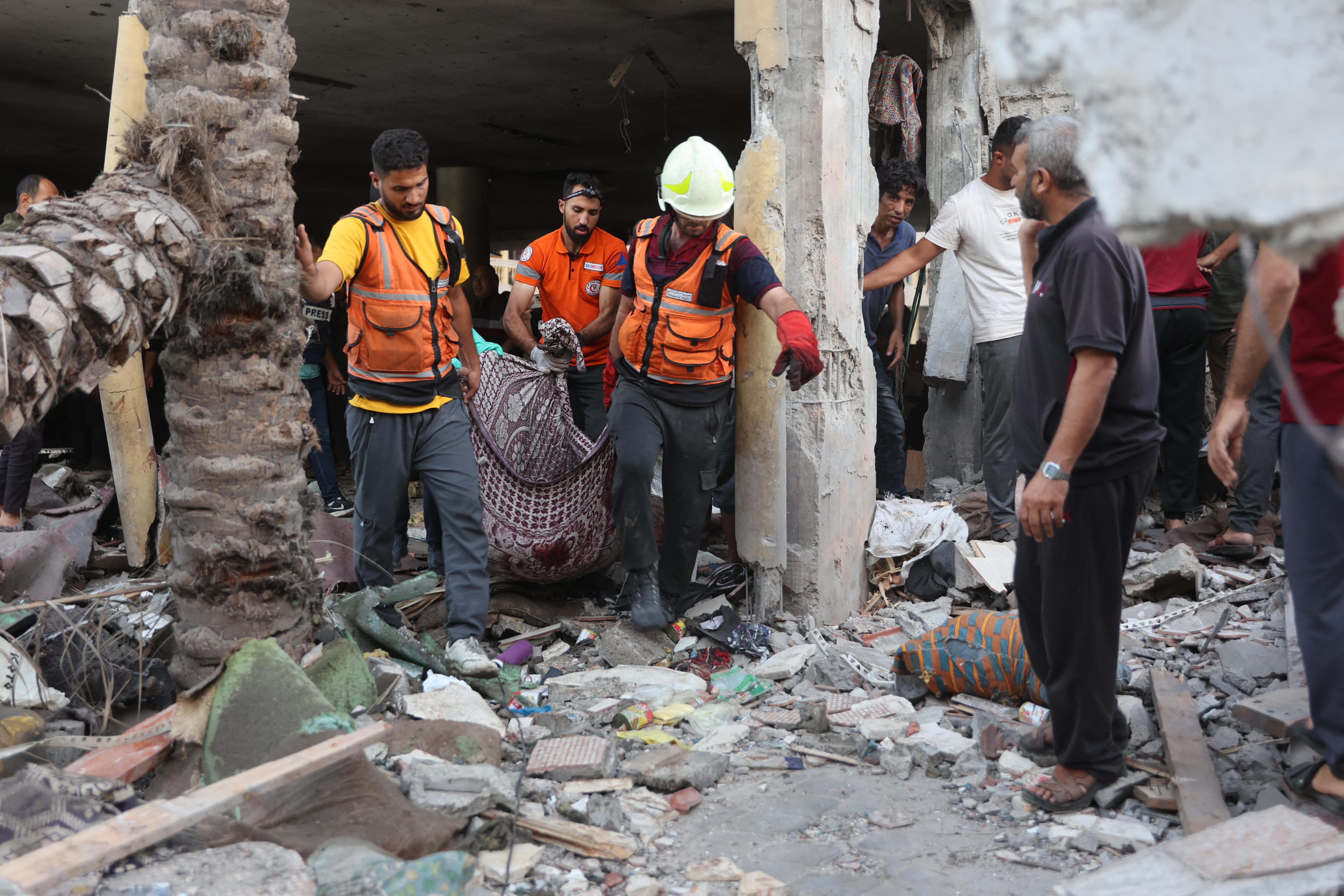Three men wearing orange carry a bag from the rubble of a building.