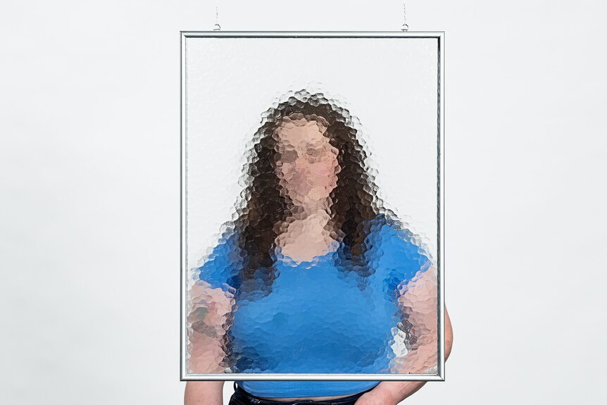 A person with long hair in a blue top stands behind a frame of glass that blurs their face.