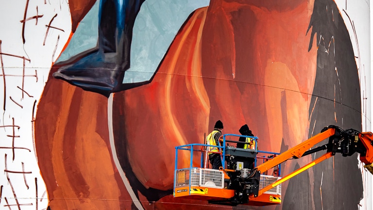 Two artists on a cherry-picker paint a large mural of a horse on a grain silo.