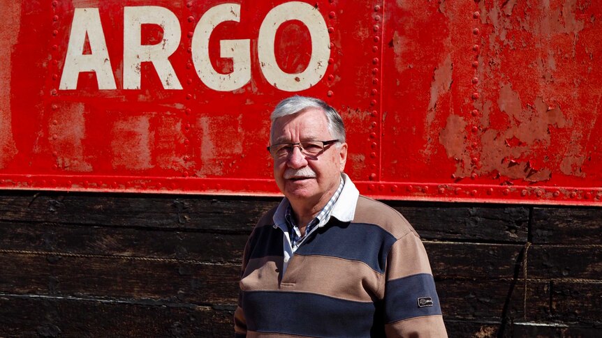 Man stands in front of the side of a barge, with flaking red paint and the word Argo in white.