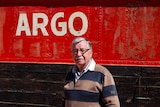 Man stands in front of the side of a barge, with flaking red paint and the word Argo in white.