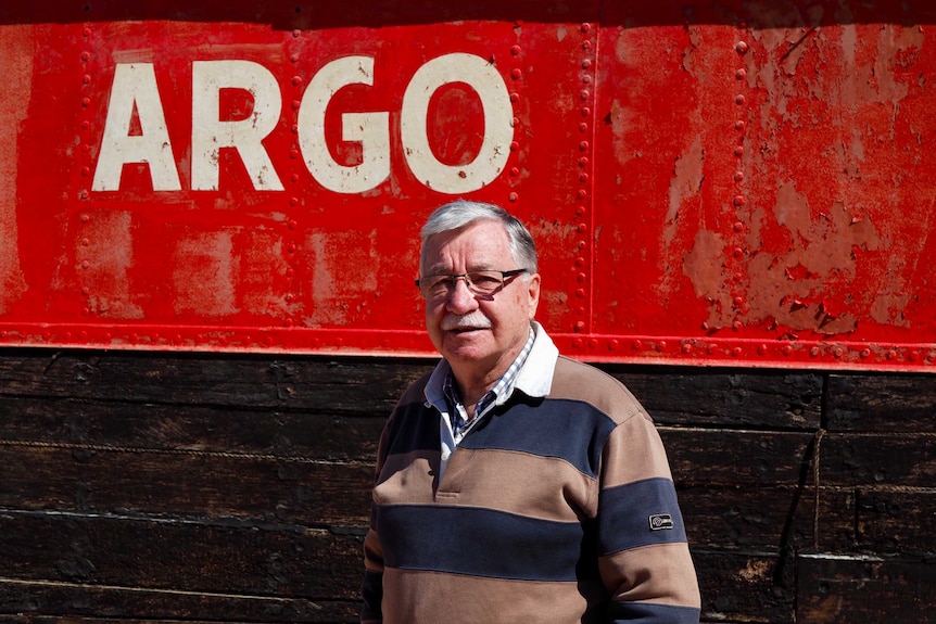 Man stands in front of the side of a barge, with flaking red paint and the word Argo in white.