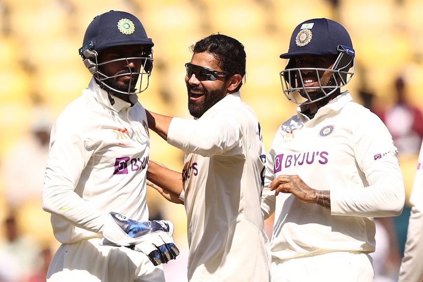 Ravindra Jadeja smiles and claps a teammate wearing a helmet on the back, as another man wearing a helmet stands beside him