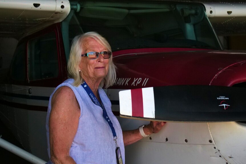 An elderly woman in a light blue shirt and glasses, with white hair, stands next to a white and red airplane inside a dark room.