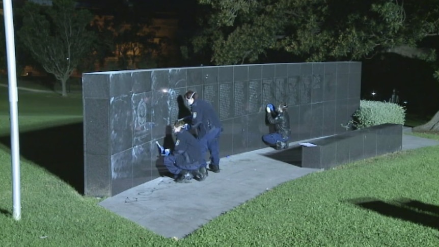 Forensic police examine the wall for fingerprints while another cleans the black graffiti off.