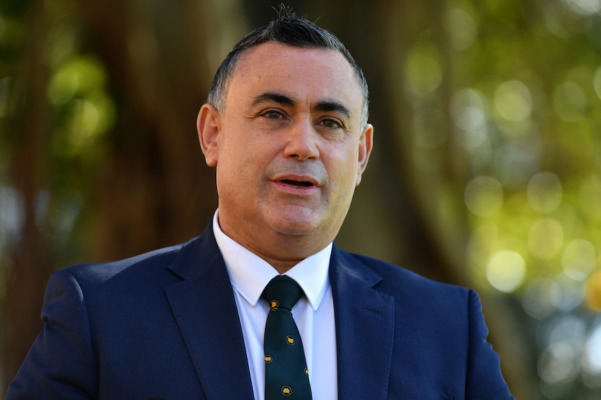 a man standing outdoors talking. He is wearing a navy blue suit and tie, with a white business shirt