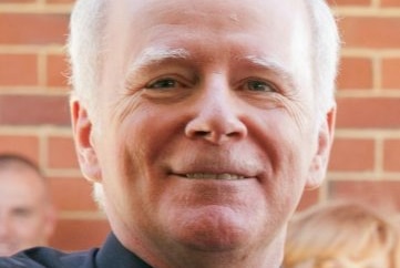 A headshot of a priest with white hair standing in front of a brick wall.