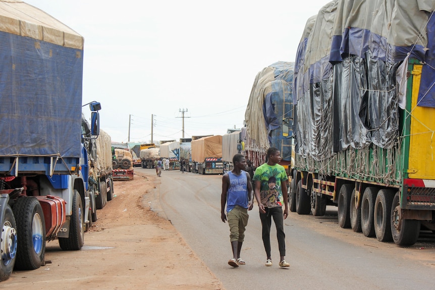 Two men walk past a long line of trucks stopped on the side of a road