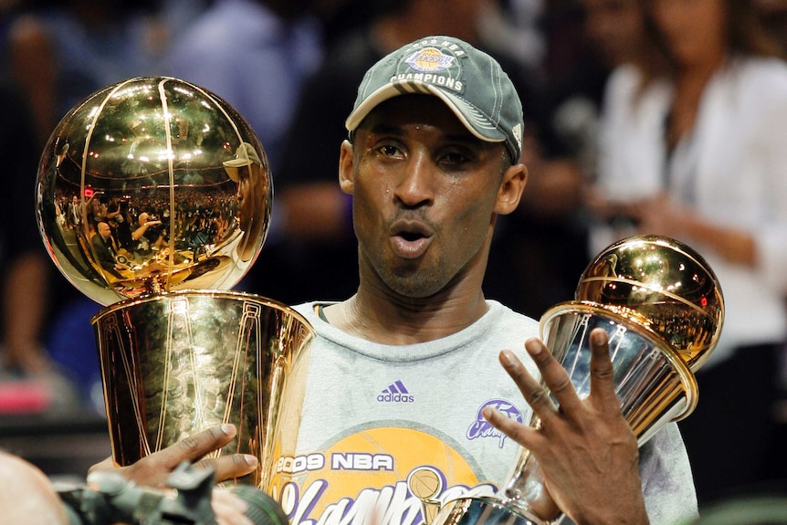 Kobe Bryant holds two trophies and holds up four fingers with his mouth open