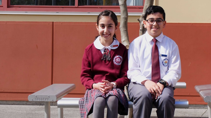 Assyrian school students Mary Anoya and Ramel Zia sit on a bench in uniforms.