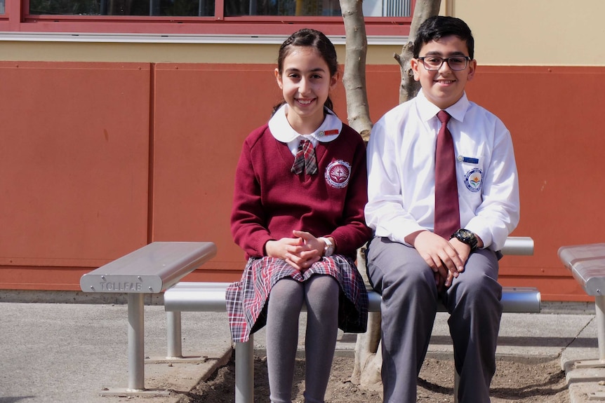 Assyrian school students Mary Anoya and Ramel Zia sit on a bench in uniforms.