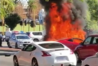 A wide shot showing a stolen Ferrari burning in the street after crashing, with a police car and other vehicles stopped nearby.