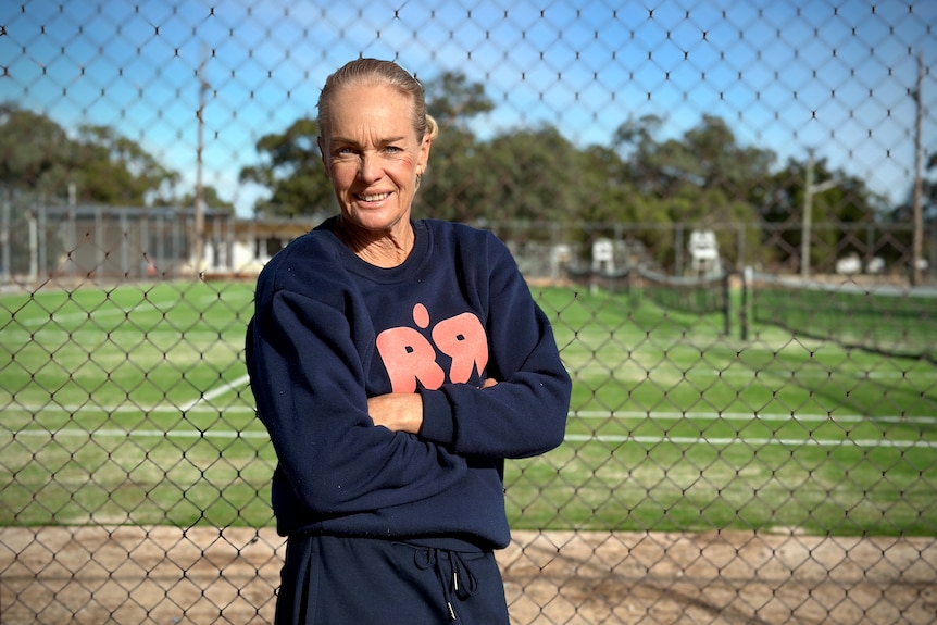 A woman in a sports jumper stands with her arms crossed looking at the camera with tennis courts behind her