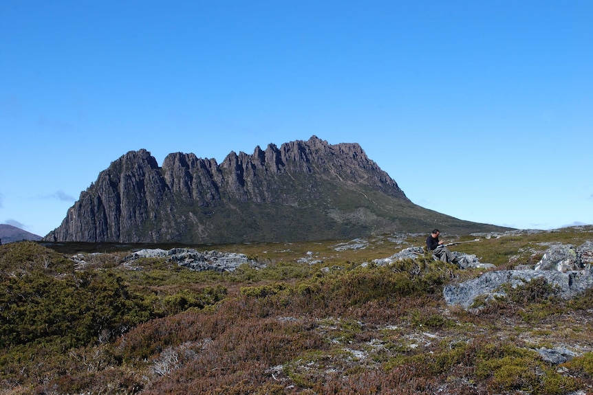 a man is sitting on a rock with a blue sky and large cliff behind him.