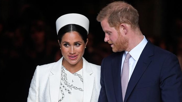 Meghan, left, wears a white dress, coat and hat and holds hands with Harry in a dark suit as they enter gate with guards by side