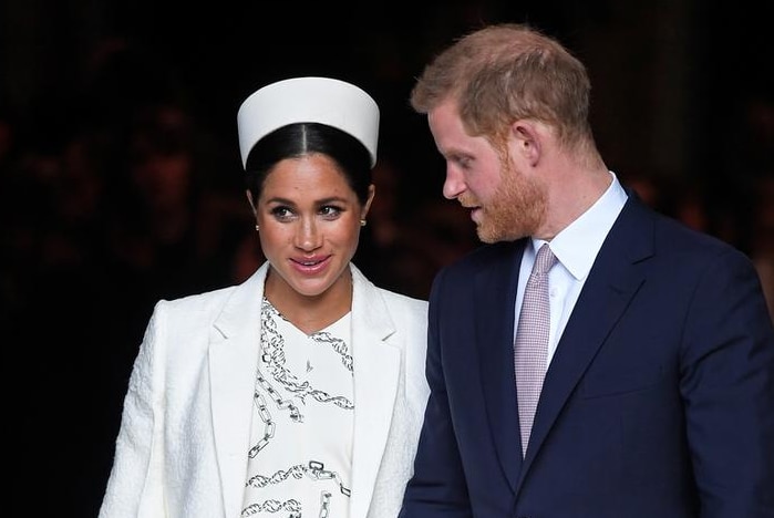 Meghan, left, wears a white dress, coat and hat and holds hands with Harry in a dark suit as they enter gate with guards by side
