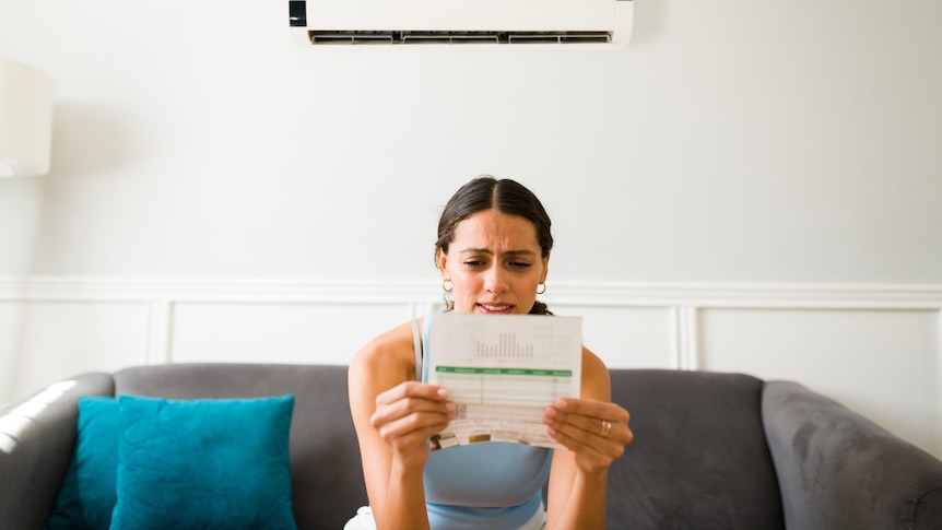A woman sitting on a couch looks at a bill with concern.